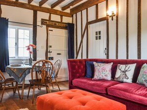 A room with exposed wooden beams, a red sofa with floral cushions, a wooden dining table with chairs, and a window with curtains. - The Cottage at Harple Farm (Detling, near Maidstone)