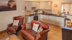 A cozy living room with two leather sofas and a dining table, a kitchen with white cabinets and a microwave, and a large window with a view of the countryside. - A relaxing, rural retreat for two. (Wrelton, near Pickering)