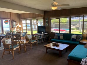 A cozy living room with a sofa, chairs, a coffee table, and a TV. Large windows offer a view of the outdoors. - Spectacular Oceanfront Puako Cottage  (Kamuela)