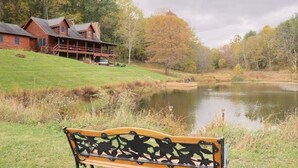 A wooden cabin with a porch, a bench in the foreground, and a pond in the background.