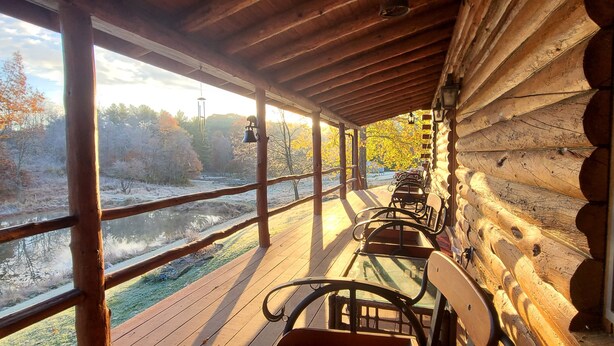 A wooden cabin with a porch overlooking a river, featuring a bench and a view of autumn trees.