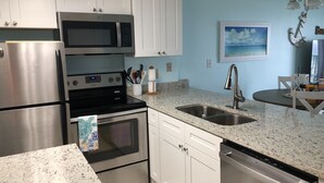 A modern kitchen with stainless steel appliances, white cabinetry, granite countertops, and a dining area with a table and chairs. - Ocean View, Beach Front Condo with Pools at Plantation Palms in Fort Morgan (Gulf Shores)