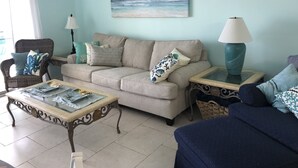 A living room with a beige sofa, a glass coffee table, and a blue armchair. There is a painting of a beach on the wall, and a lamp on a side table. - Ocean View, Beach Front Condo with Pools at Plantation Palms in Fort Morgan (Gulf Shores)