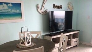 A dining area with a round table, two chairs, a TV on a wooden stand, and a painting of a beach scene. - Ocean View, Beach Front Condo with Pools at Plantation Palms in Fort Morgan (Gulf Shores)