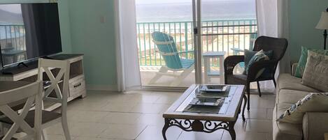 A living room with a TV, a dining table, a sofa, and a view of the beach.