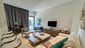 Ein modernes Wohnzimmer mit einer Couch, zwei Sesseln, einem Couchtisch und einem Fernseher. Es gibt einen Essbereich mit Tisch und Stühlen sowie einen Balkon mit Vorhängen.