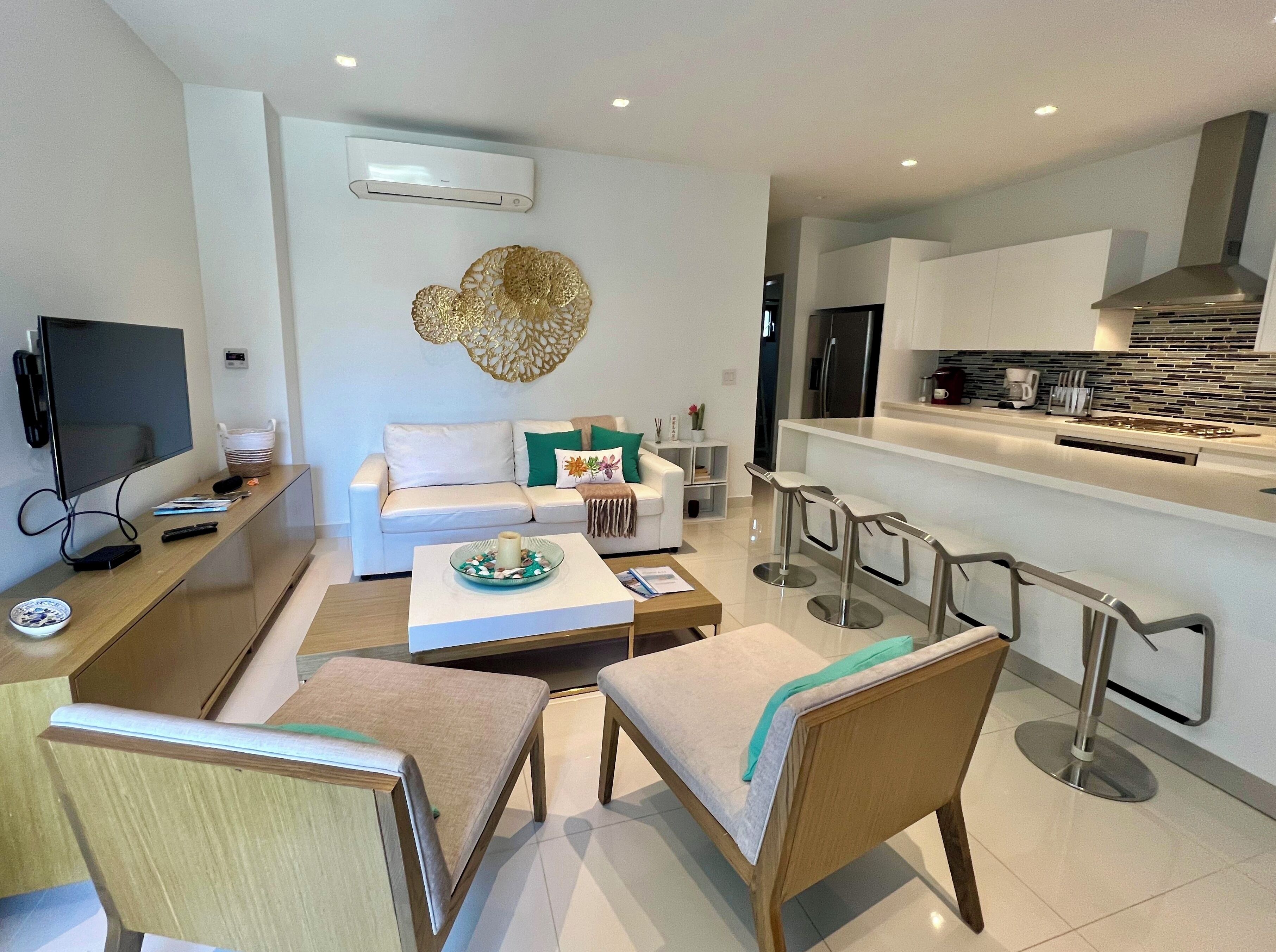 A modern living room with a sofa, coffee table, and TV. There is a kitchen area with a bar and stools.