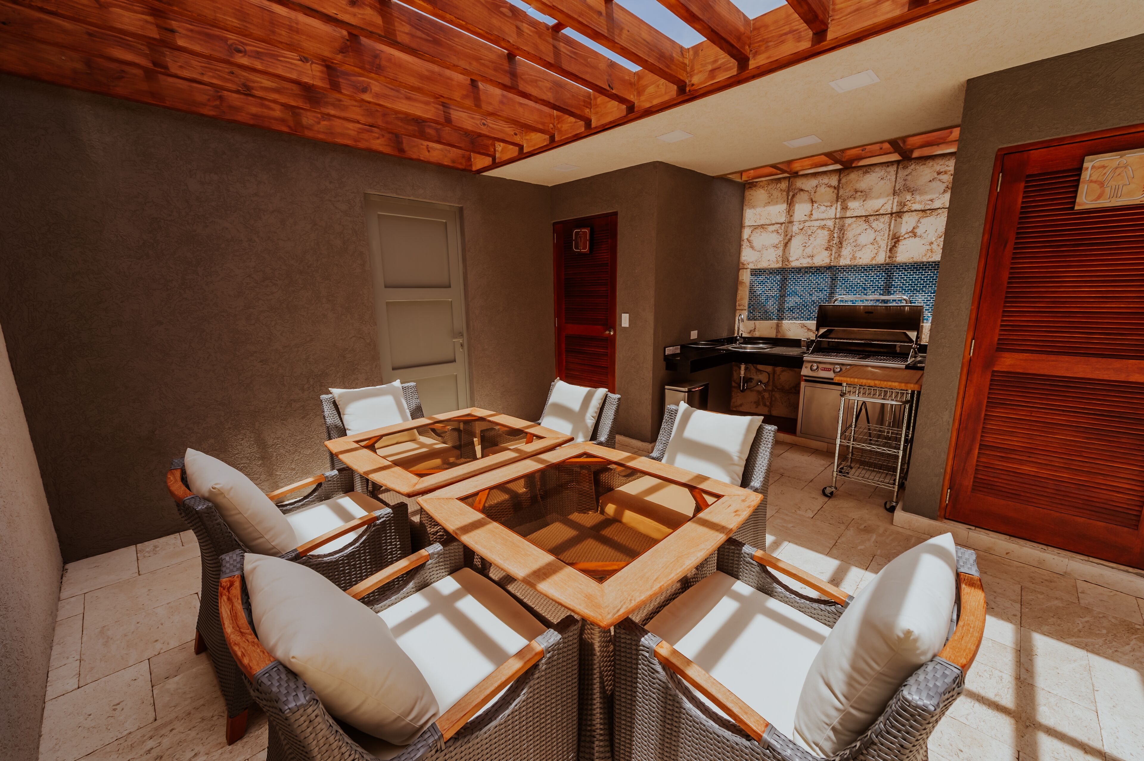 A patio with a wooden table and wicker chairs, a grill, and a tiled backsplash.