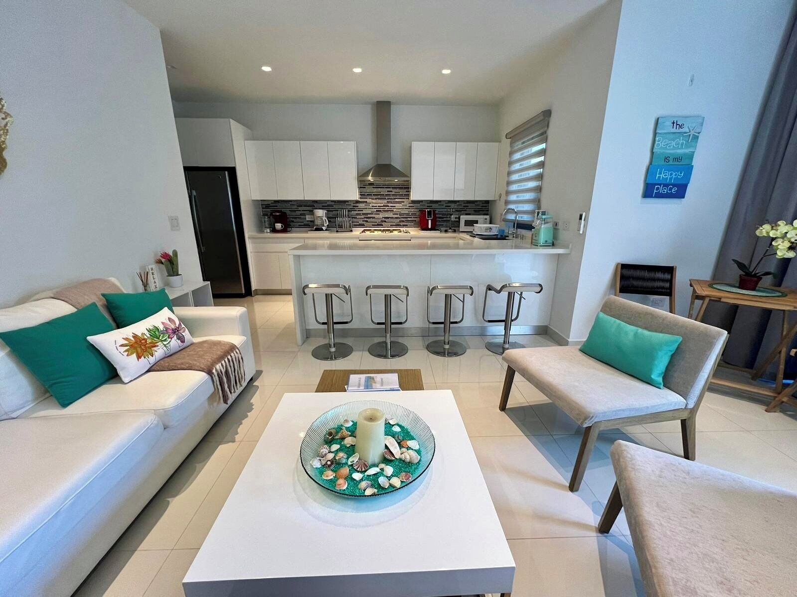 Una sala de estar moderna con un sofá blanco, dos sillones, una mesa de centro con un adorno decorativo, y una cocina al fondo con armarios blancos y una isla central con taburetes de bar.
