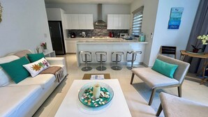Ein modernes Wohnzimmer mit einem weißen Sofa, zwei Sesseln, einem Couchtisch mit dekorativer Dekoration und einer Küche im Hintergrund mit weißen Schränken und einer zentralen Kochinsel mit Barhocker.