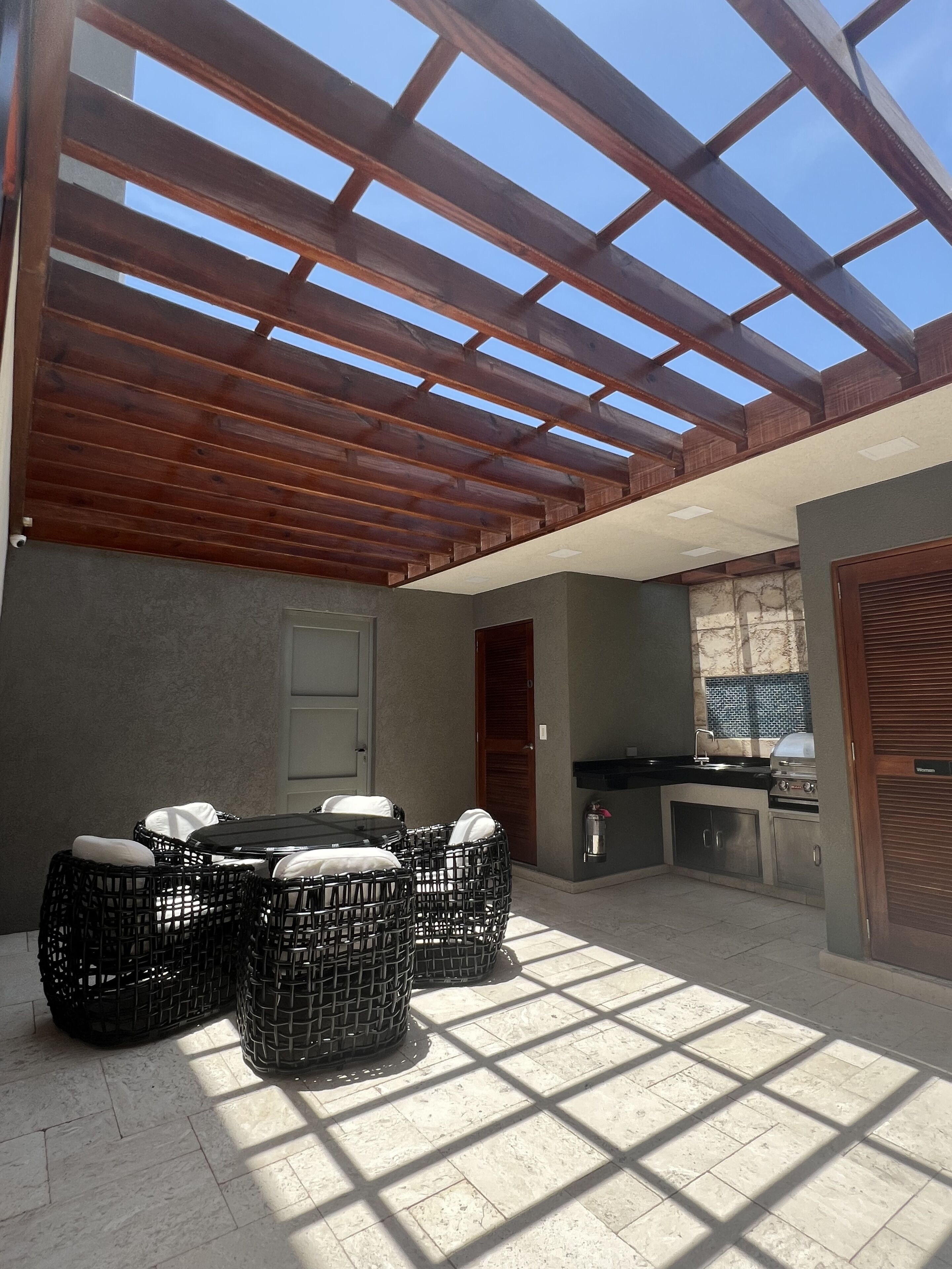 Een terras buiten met een houten pergola, een eettafel met rieten stoelen en een ingebouwde barbecue.