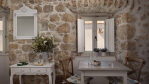 Una stanza dalle pareti in pietra con un tavolo e sedie bianche, uno specchio e una finestra con persiane.
