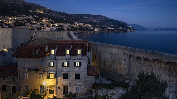 Un edificio in pietra con numerose finestre e un tetto in tegole rosse, situato accanto a un muro fortificato e una distesa d'acqua.