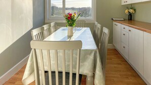 Un comedor con una mesa, sillas, un florero con flores, un ventanal y una cocina con armarios.