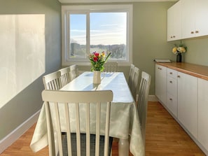 Un comedor con una mesa, sillas, un florero con flores, un ventanal y una cocina con armarios.