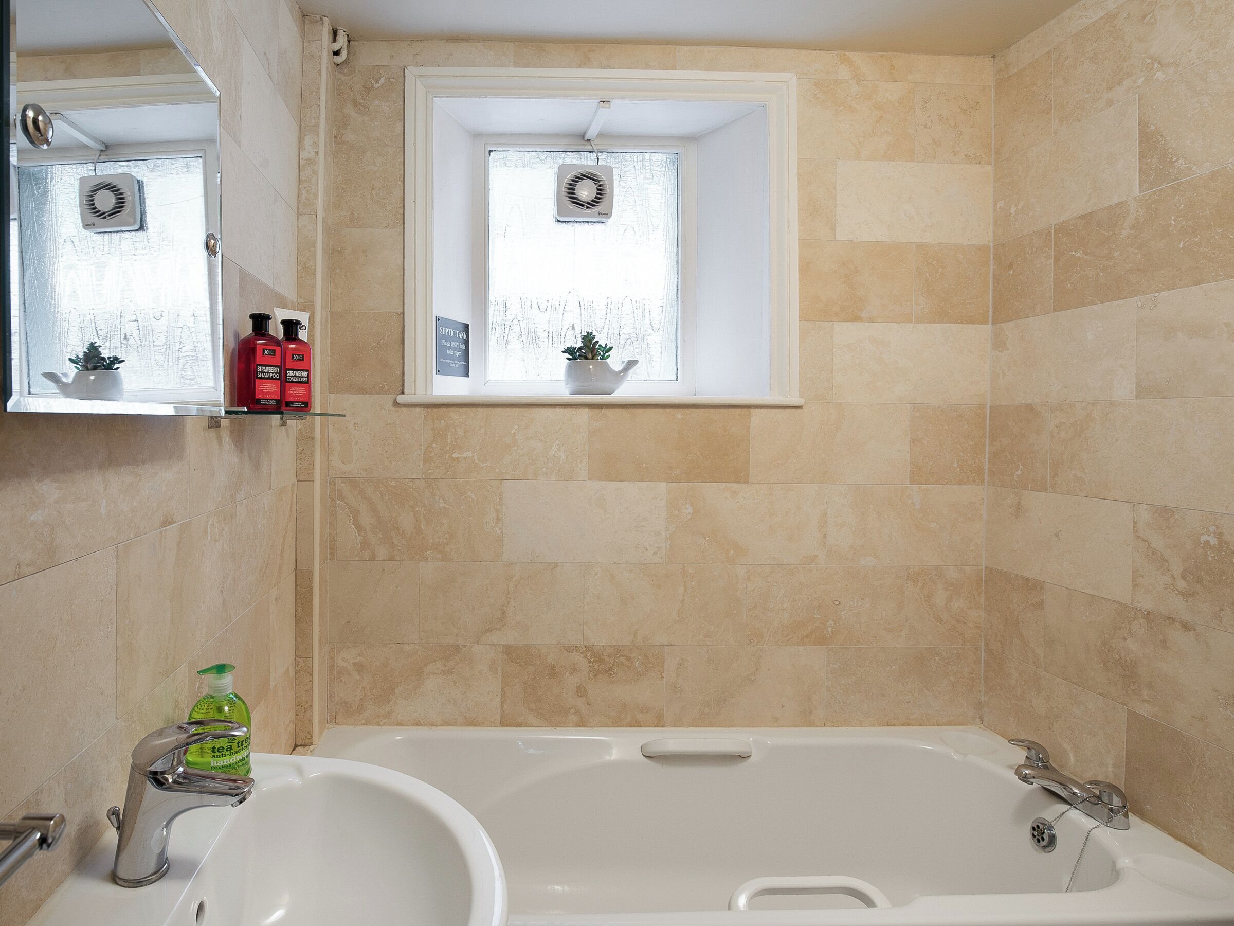 Un baño con bañera, lavamanos, ventana con ventilador y repisas con artículos de aseo.