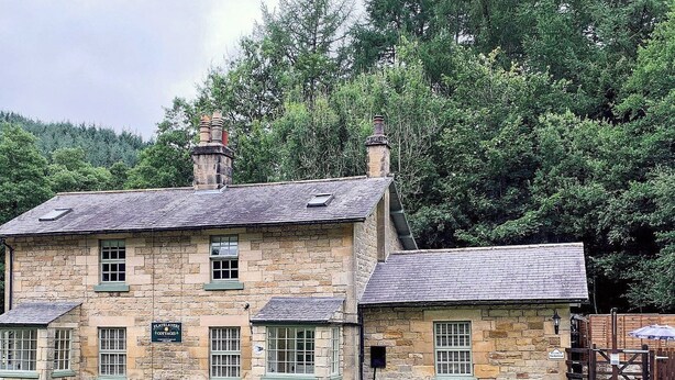 Un bâtiment en pierre doté de nombreuses fenêtres et d’une cheminée, entouré de verdure.