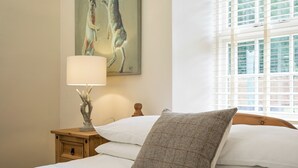 Un lit bien fait, avec du linge de lit blanc, un oreiller gris texturé, une table de chevet en bois avec une lampe, et un tableau représentant deux lapins accroché au mur.