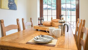 Une table de salle à manger en bois, avec des chaises, une cafetière, du pain, de la confiture et un tableau accroché au mur.