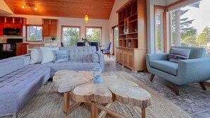 Ein großzügiger Wohnbereich mit einer großen Couch, einem Couchtisch und einem Bücherregal. Es gibt eine Küchenzeile mit Holzschränken und einer Mikrowelle. Das Zimmer verfügt über große Fenster, die natürliches Licht hereinlassen.