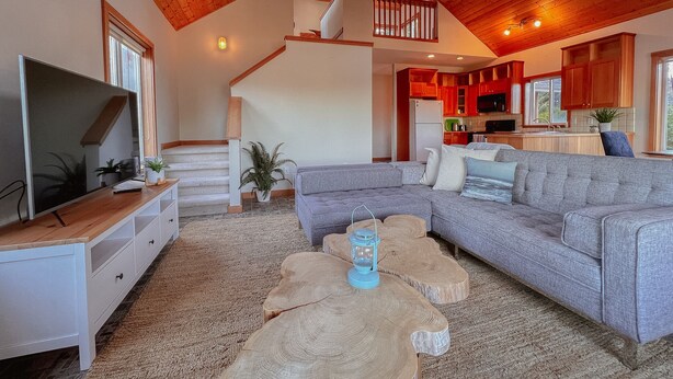 Ein großzügiger Wohnraum mit grauem Sofa, Couchtisch aus Holz und Fernseher auf einem weißen Schrank. Eine Treppe führt zu einer Küchenzeile mit roten Schränken.