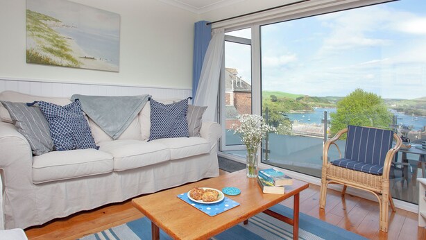 Ein Wohnzimmer mit einer Couch, einem Holztisch, einem Korbstuhl und einem großen Fenster mit Blick auf das Meer und Gebäude.