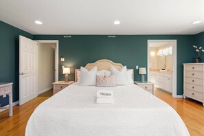 A bedroom with a large bed, two bedside tables, a dresser, and a door leading to another room.