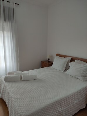 A neatly made bed with white linens and pillows, a wooden nightstand with a lamp, and sheer curtains on the window.