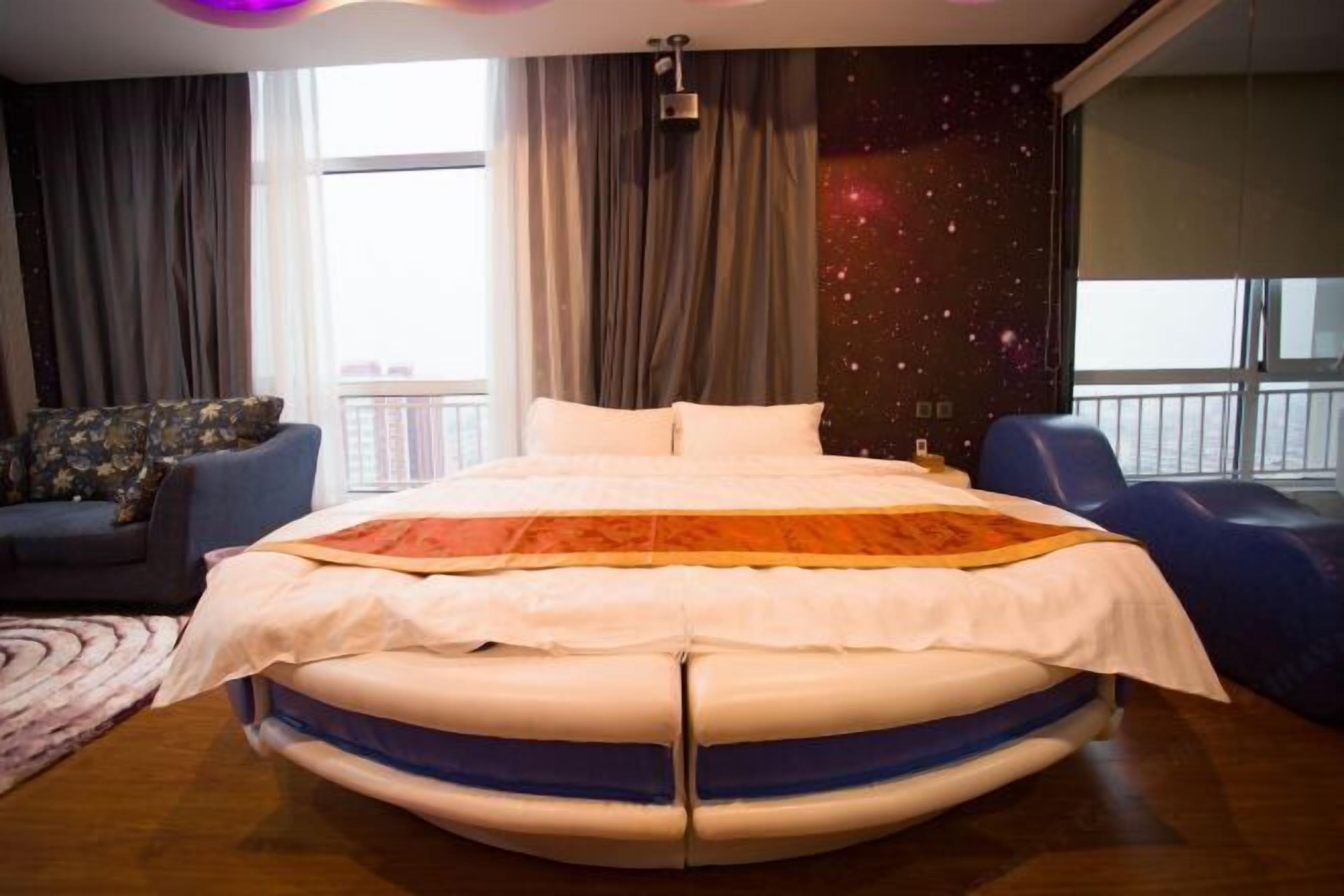 A modern bedroom with a circular bed, a view of the city, and a galaxy-themed wall.