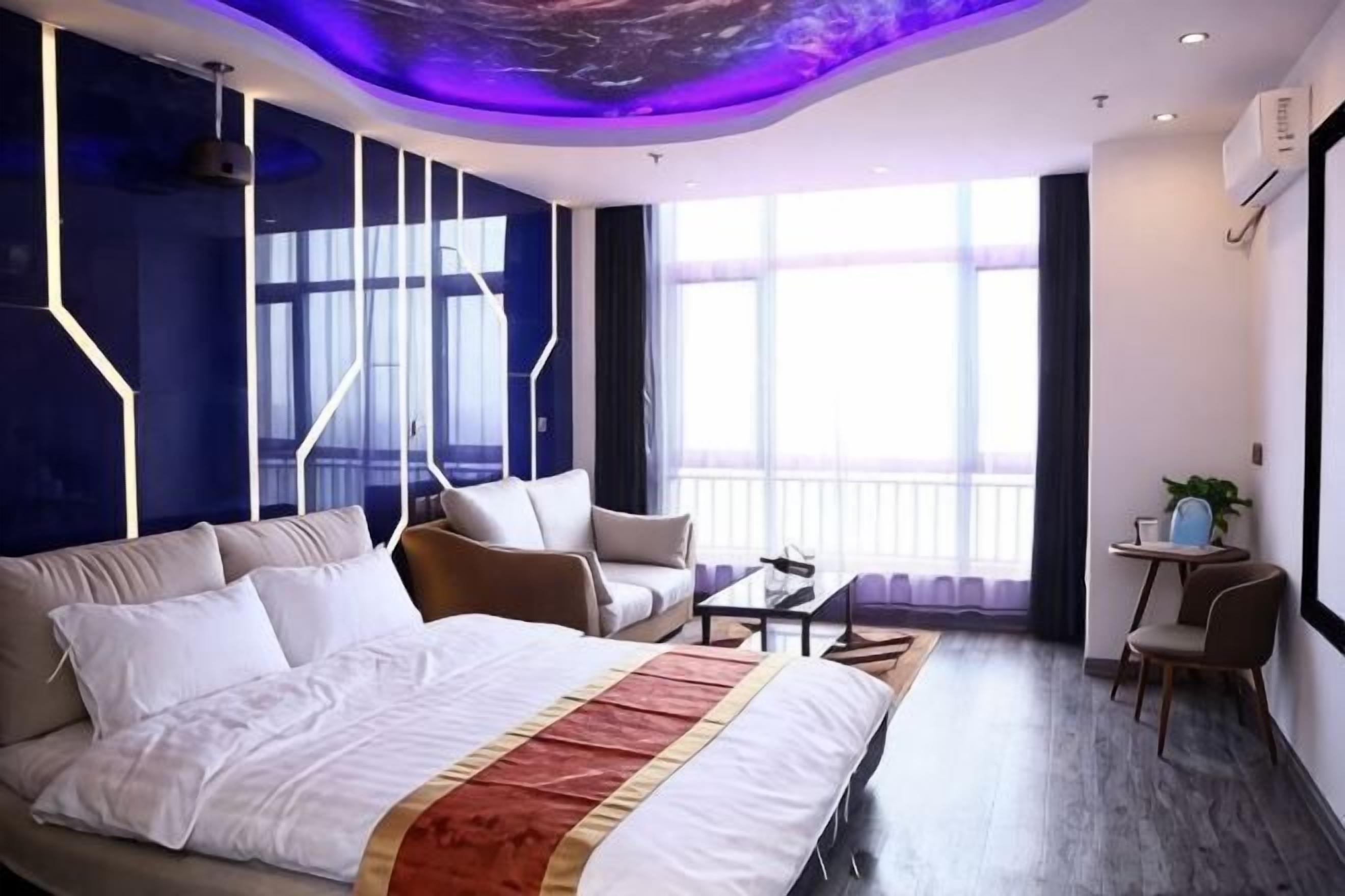 A modern hotel room with a large bed, a sofa, a small table, and a chair. There is a window with a view of the outside.
