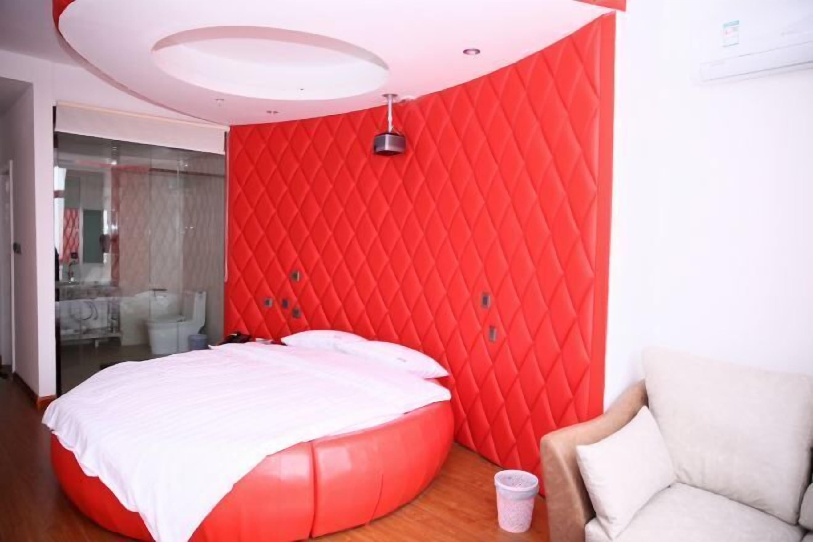 A modern bedroom with a red quilted headboard, a round bed with white bedding, a beige armchair, and a glass shower enclosure.