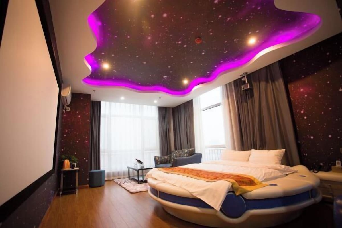 Un dormitorio moderno con una cama curvada, un techo con temática espacial y un ventanal con cortinas.