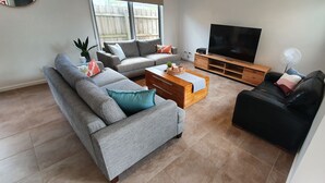 Un salon avec un canapé gris, un canapé en cuir noir, une table basse en bois, un téléviseur posé sur un meuble en bois et un ventilateur.