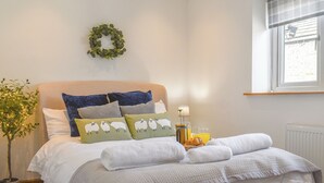 A neatly made bed with white linens, green and blue pillows, a small table with a lamp and breakfast items, a potted plant, and a window with blinds. - 3 bedroom accommodation in Skipton (Skipton)