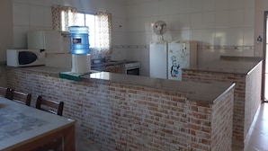 A kitchen with a brick counter, a microwave, a water dispenser, a refrigerator with a penguin decoration, and a window with patterned curtains. - Country house in Araçoiaba da Serra (Araçoiaba da Serra)