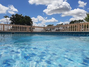 Une piscine d’un bleu éclatant, bordée de carreaux, entourée d’une balustrade beige et d’une rangée de chaises longues noires.