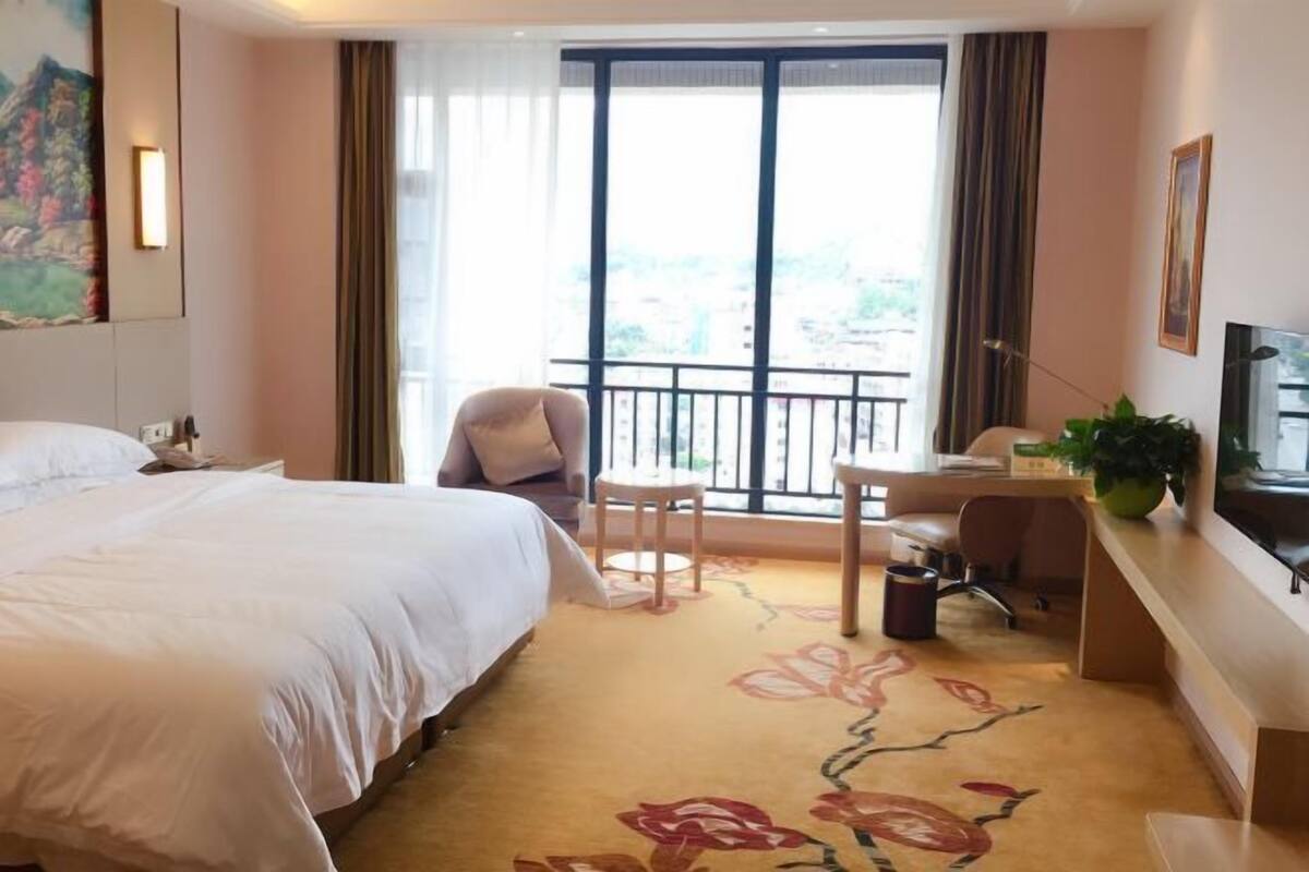 Habitación de hotel con cama, escritorio, silla, televisor y balcón con cortinas.
