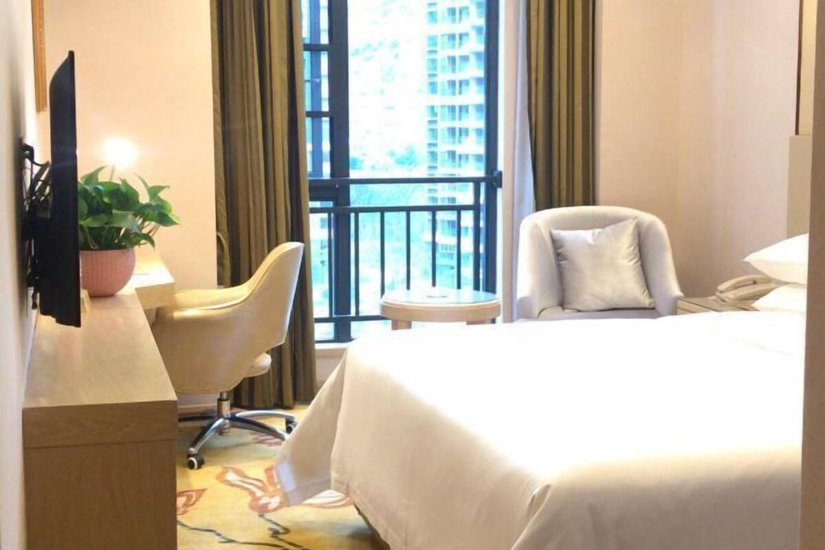 Habitación de hotel con cama, escritorio, silla, una mesita, televisor y ventana con vistas a edificios.