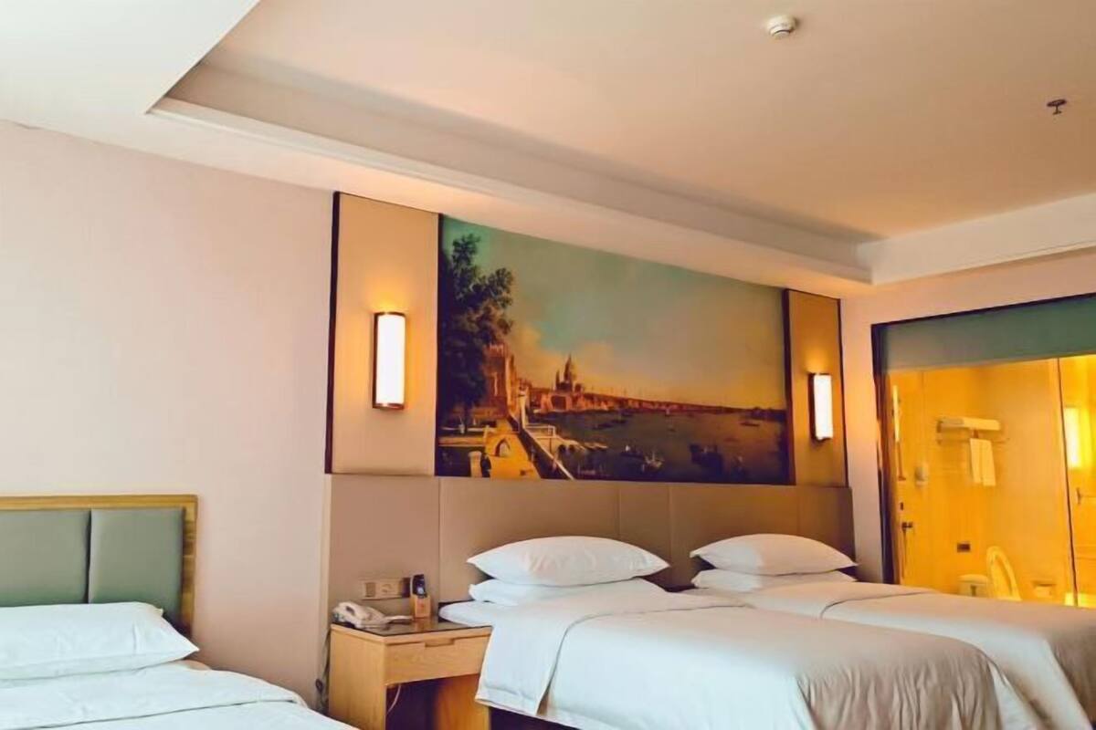 Habitación de hotel con dos camas, una mesita de noche, un cuadro de un paisaje urbano y una zona de ducha con cerramiento de vidrio.