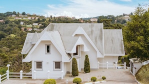 A white two-story house with a steeply pitched roof, a white fence, and a paved driveway.