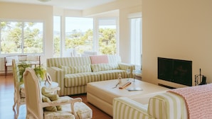 A living room with a sofa, armchairs, a coffee table, and a television. There are large windows providing a view of trees and hills.