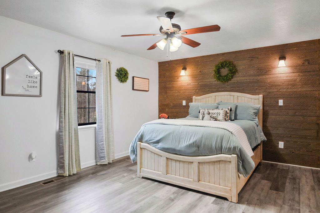 A bedroom with a wooden wall, a bed with a headboard, a ceiling fan, and a window with curtains.