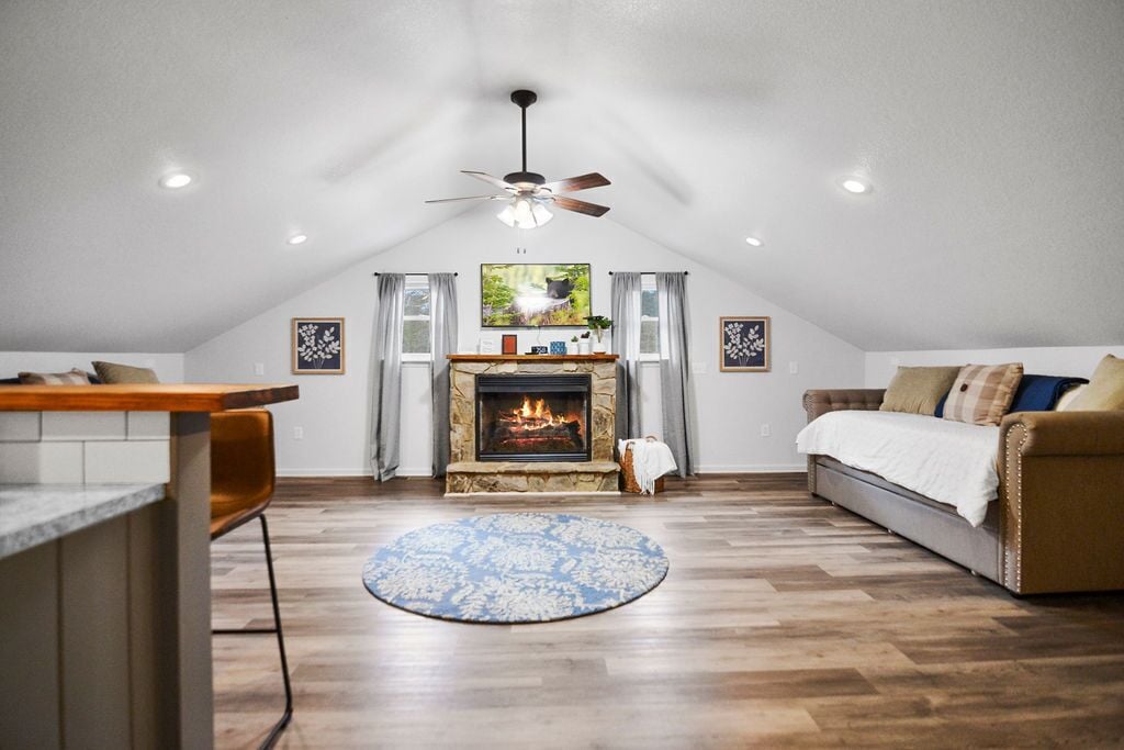 A cozy loft bedroom with a fireplace, a ceiling fan, and a large window.