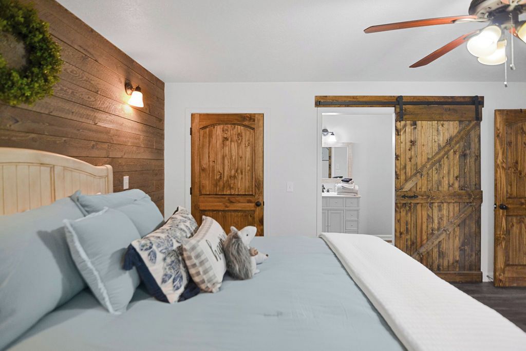 Une chambre avec une tête de lit en bois, un lit avec un matelas blanc et un ventilateur de plafond. On aperçoit deux portes en bois et une porte coulissante de style rustique au fond.