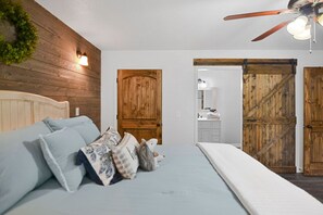 Une chambre avec une tête de lit en bois, un lit avec un matelas blanc et un ventilateur de plafond. On aperçoit deux portes en bois et une porte coulissante de style rustique au fond.