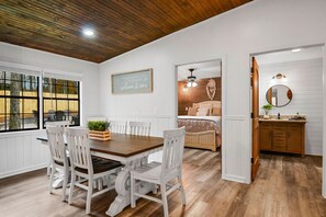 Un coin repas avec une table en bois et des chaises blanches, une cuisine avec des meubles en bois et une chambre avec un lit et un attrape-rêves suspendu.