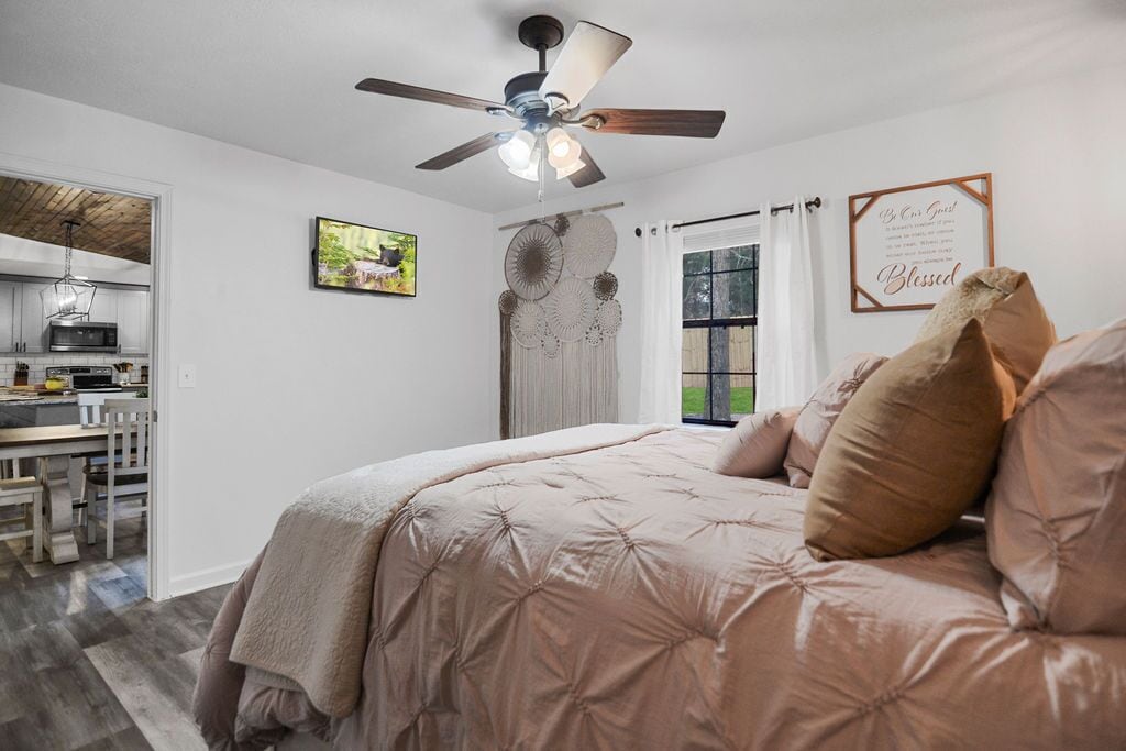 A bedroom with a bed, a ceiling fan, a window with curtains, and a kitchen visible through an open door.