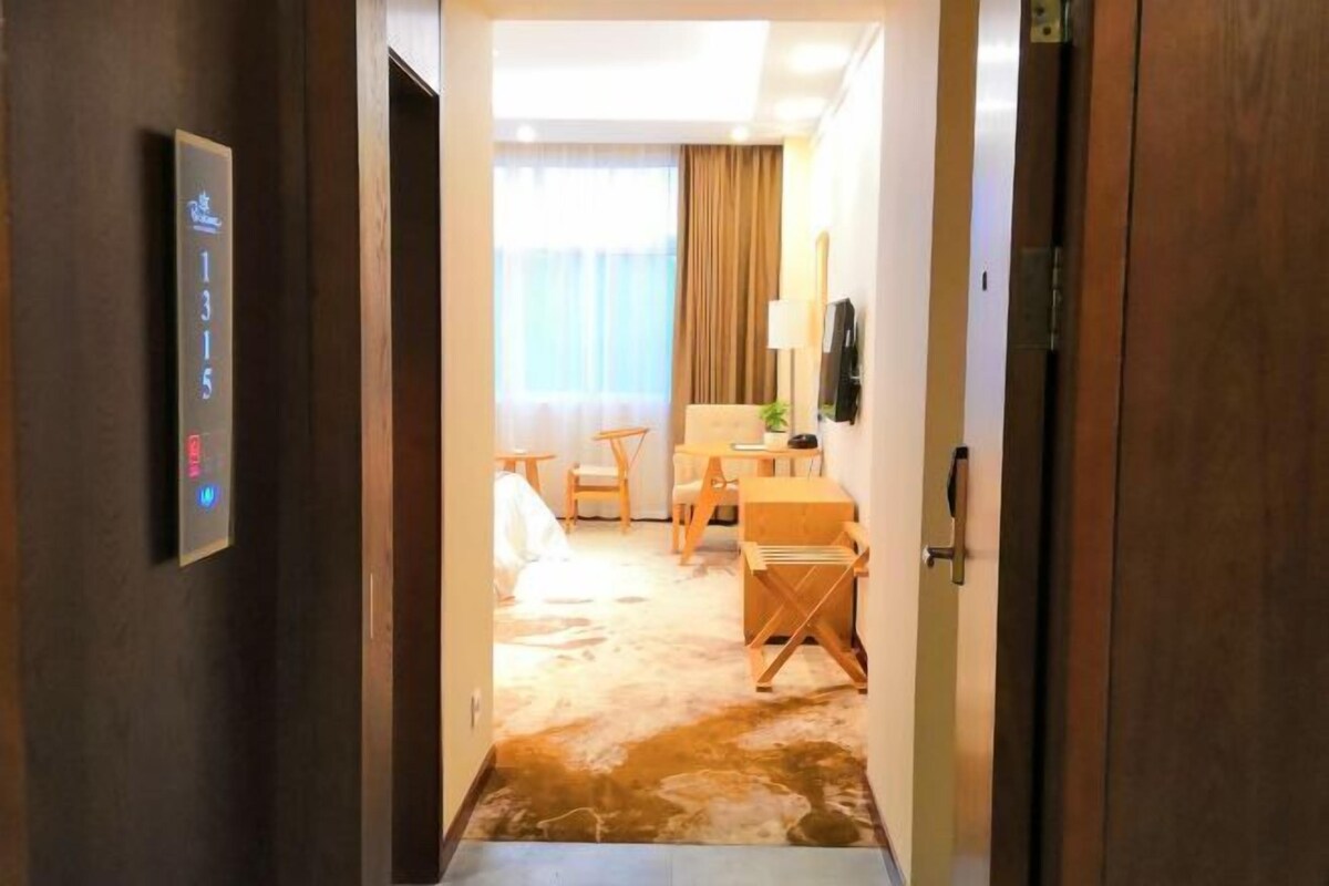 Quarto de hotel com porta de madeira, painel de controle, cama, escrivaninha, cadeira e uma janela grande com cortinas.