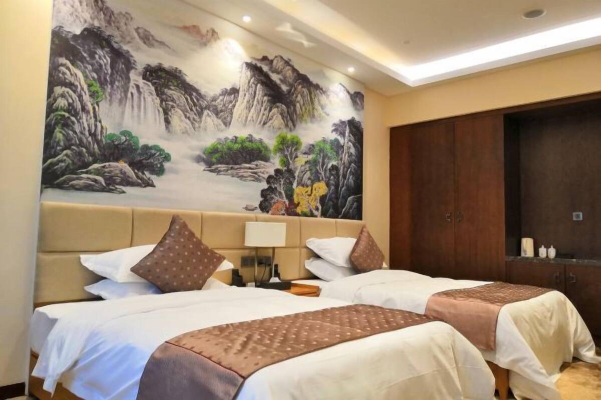 Een hotelkamer met twee bedden, een groot schilderij van een berglandschap aan de muur, een houten garderobe en een nachtkastje met een lamp.