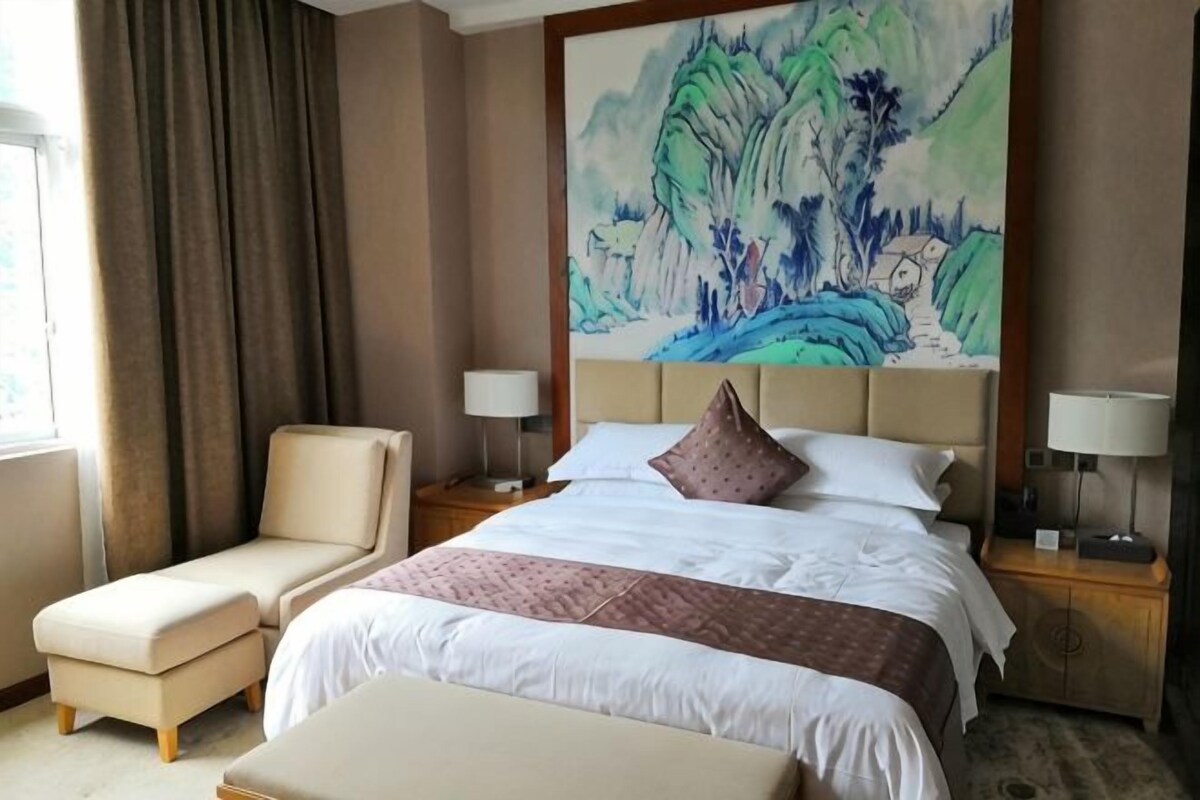 Hotellihuone, jossa on suuri sänky, tuoli, työpöytä ja taulu seinällä.